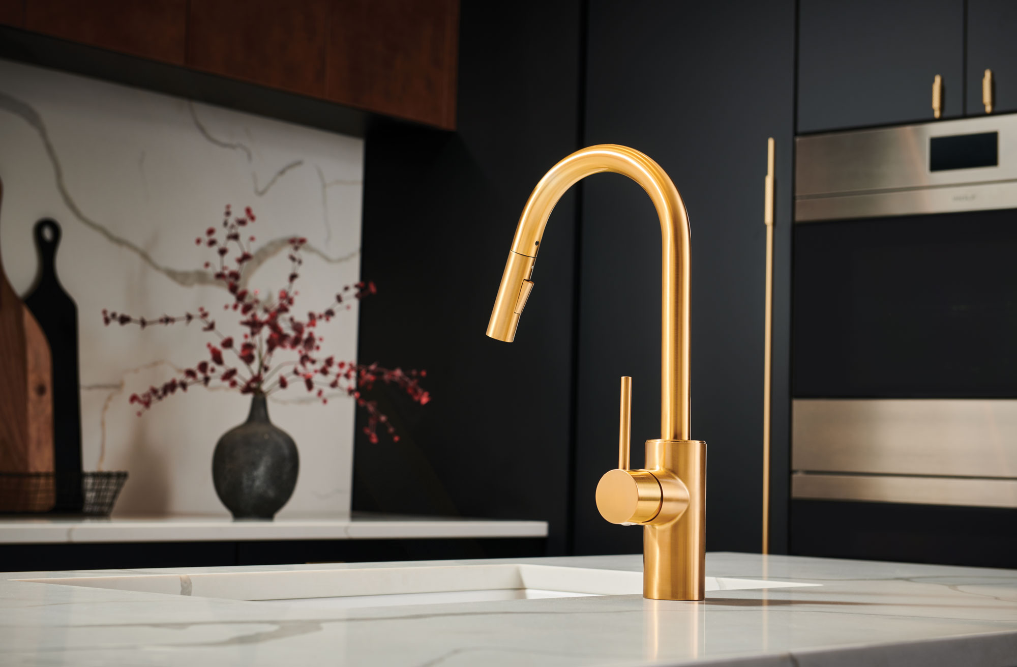 A sleek gold kitchen faucet on a marble island, surrounded by dark cabinetry and stainless steel appliances. A modern vase with red branches adds elegance.