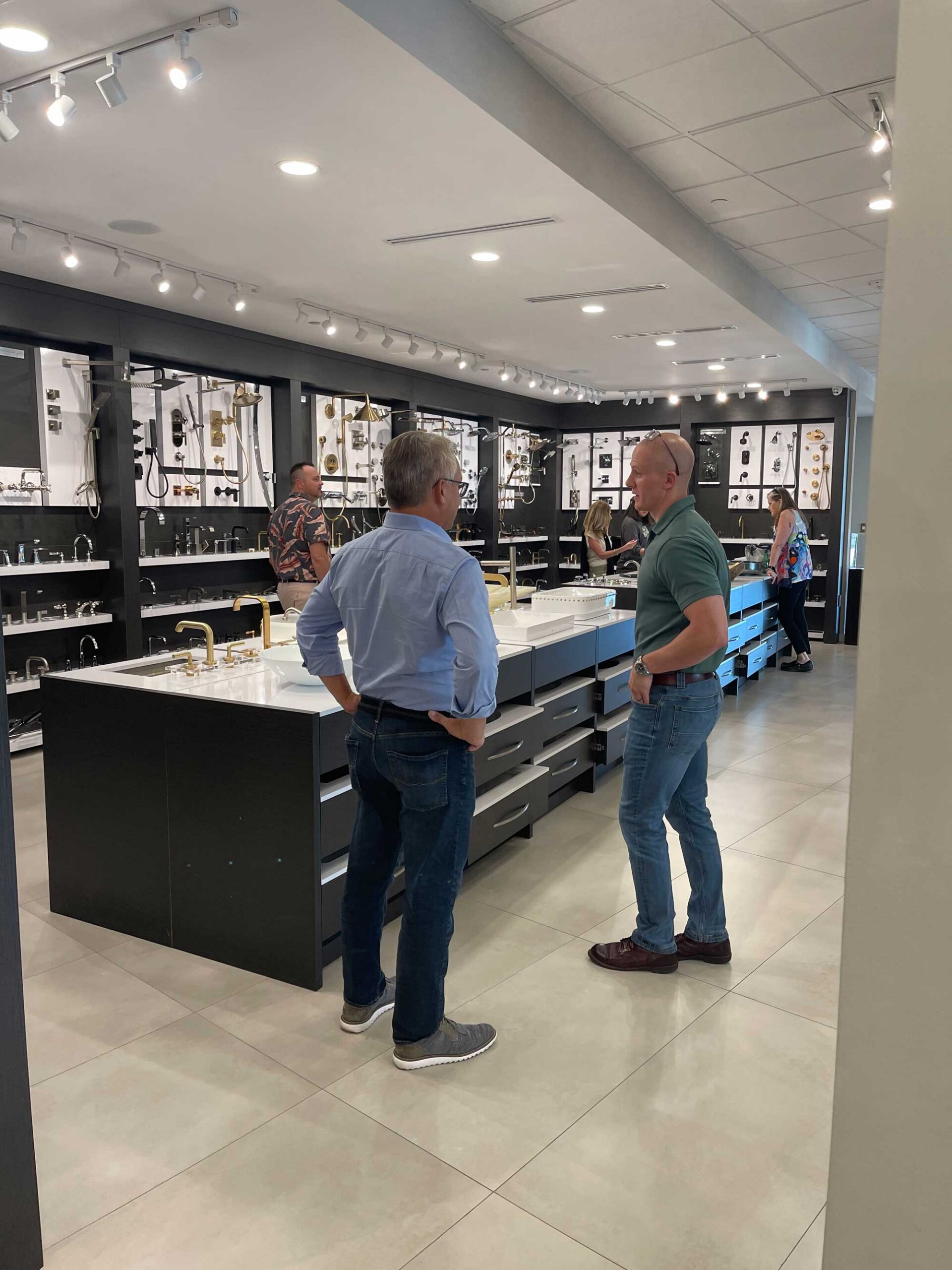 4CBD979A-8A2F-47C9-A49D-A7263B487151 Two men stand in a store, beside a counter, with shelves filled with products in the background.