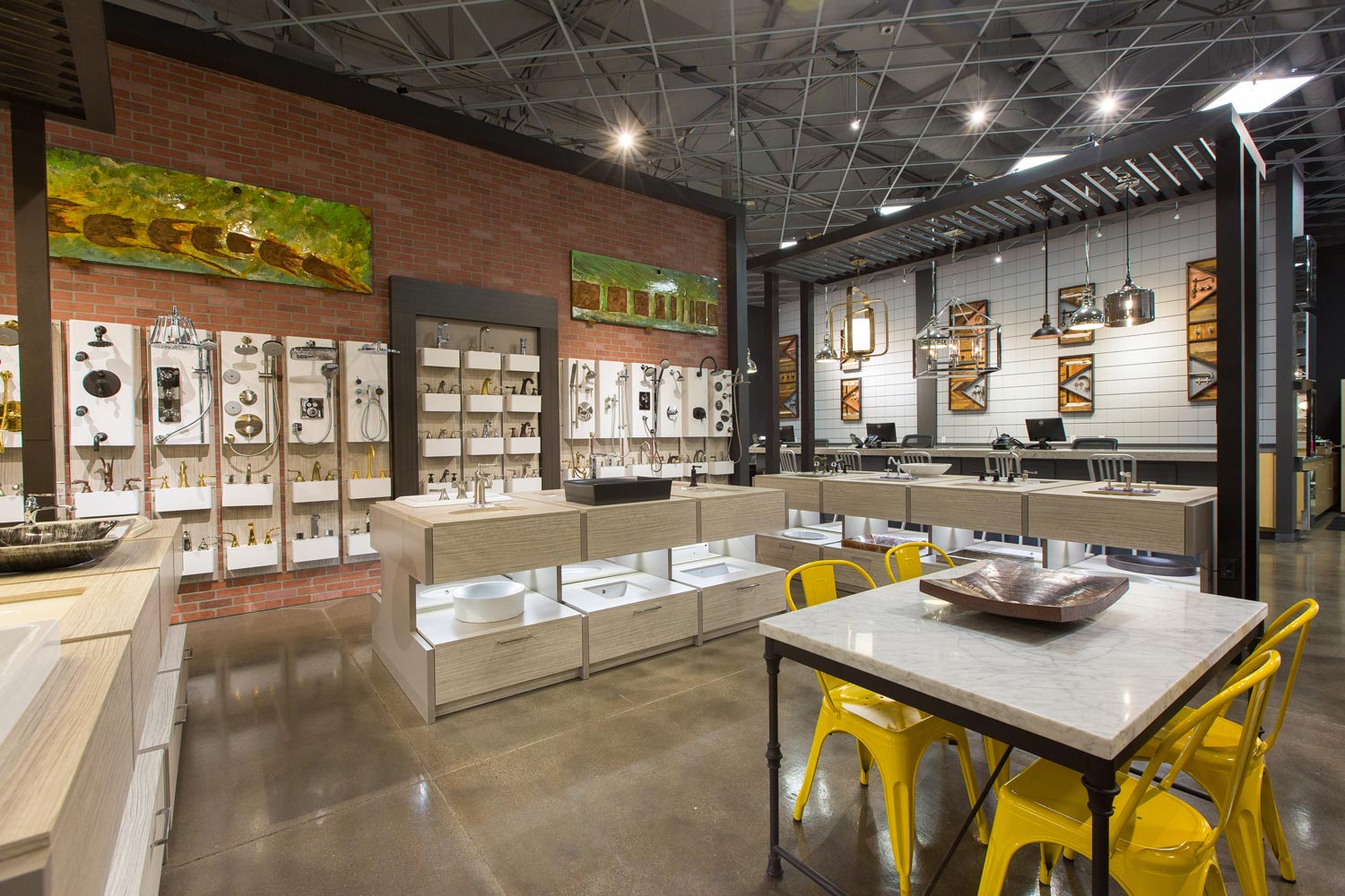 Modern showroom with faucets and sinks on display under industrial lighting. A brick accent wall, yellow chairs, and sleek fixtures create a contemporary vibe.