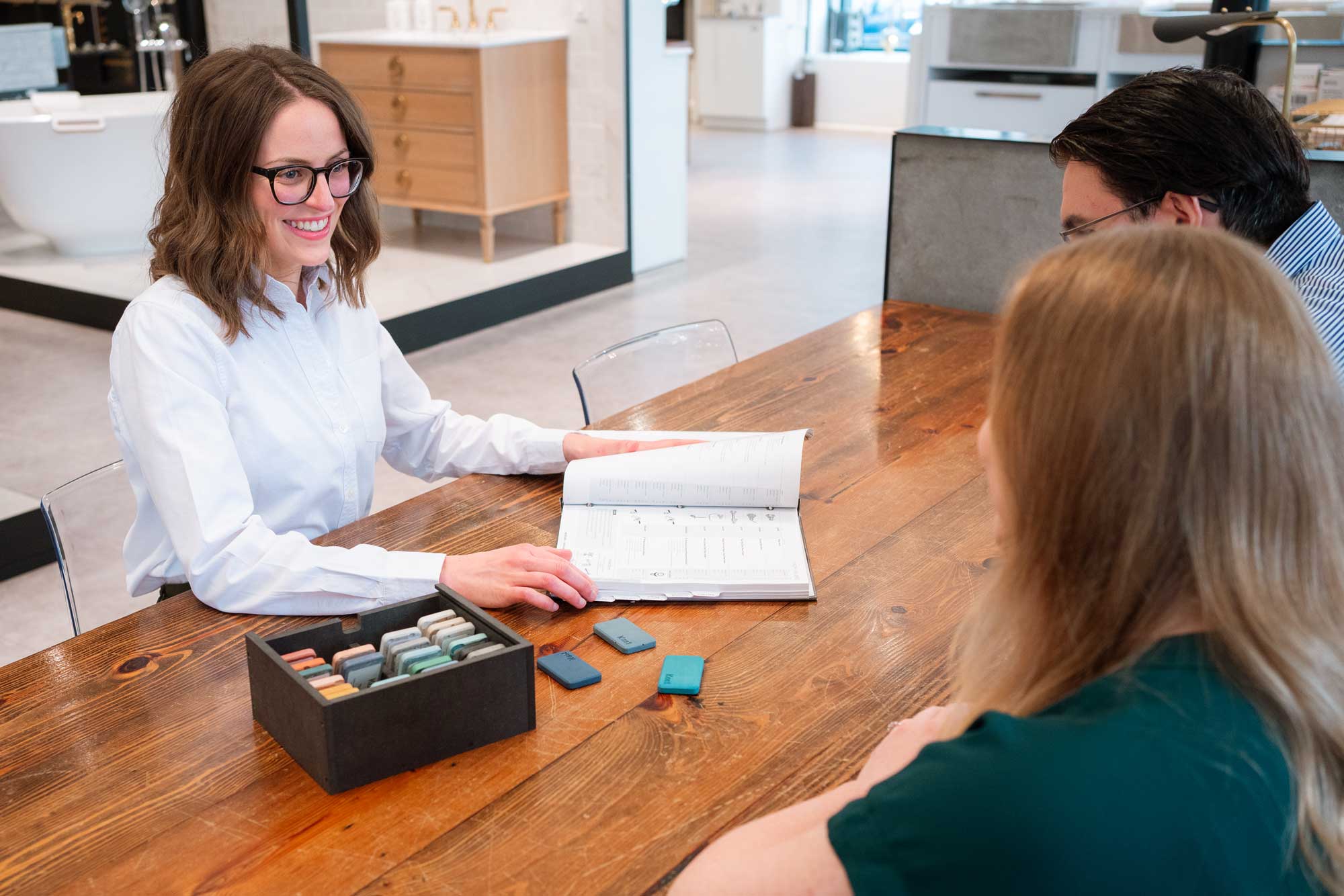 A woman shows a catalog to two people at a wooden table, with a box of sample materials and several samples spread out in front of them.