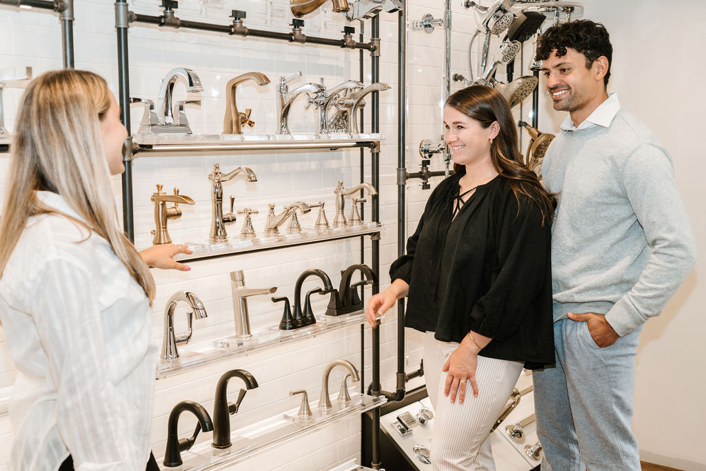 A couple smiles and listens to a showroom employee while browsing various metallic faucets displayed on shelves. The atmosphere is bright and engaging.