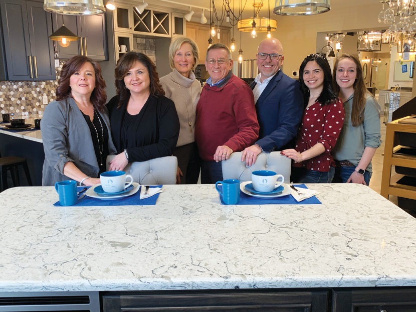 A group of seven people smiling, standing behind a kitchen island with white marble countertop. Blue mugs and saucers are set on the island. Warm, inviting atmosphere.